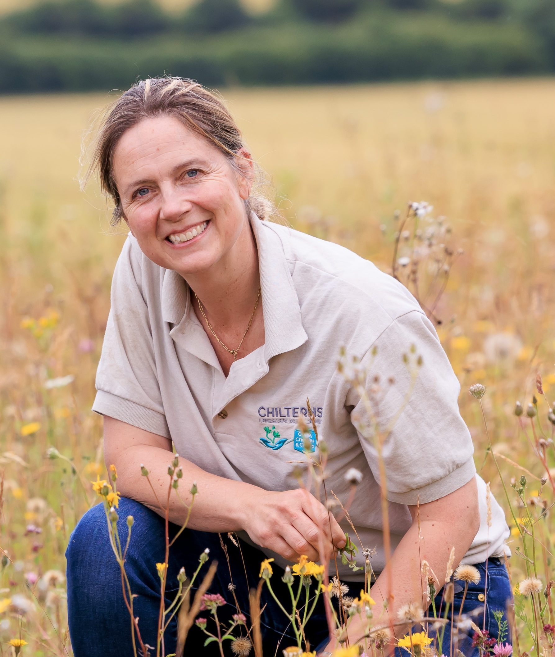 A woman wearing a beige polo shirt and jeans kneels in a wildflower meadow.