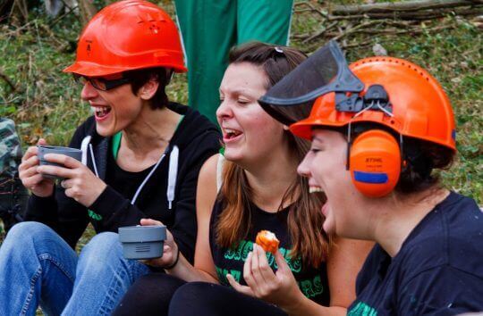 Three female volunteers sit and have tea and snacks outdoors, two of them wearing hard hats