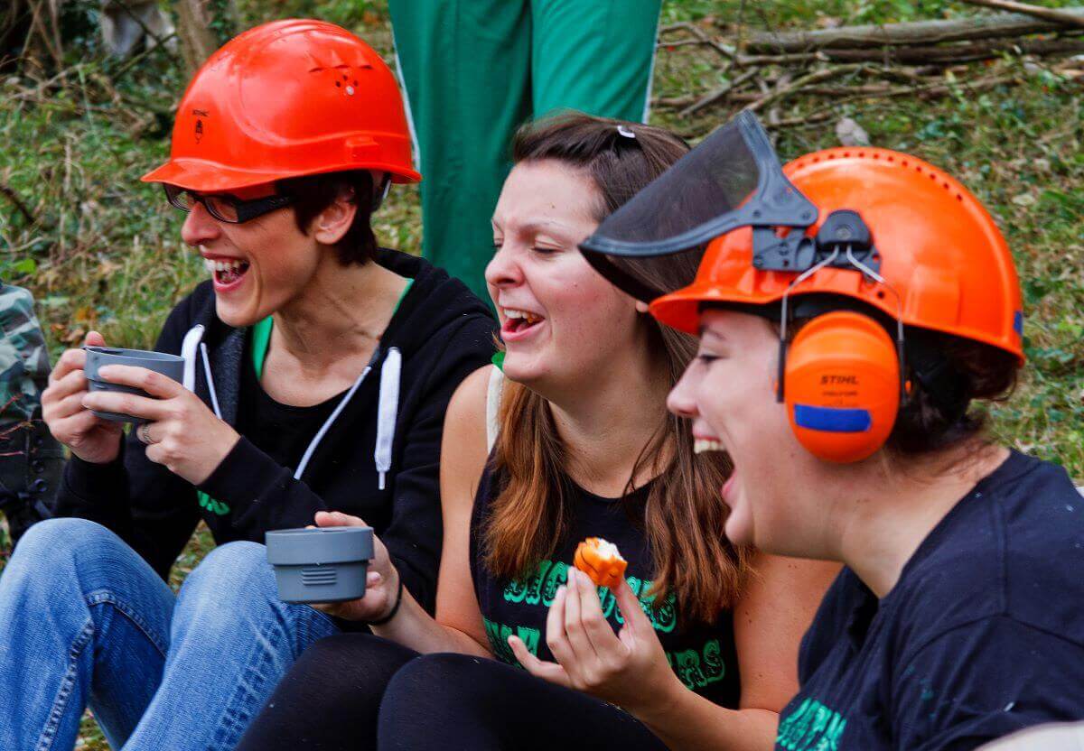 Three female volunteers sit and have tea and snacks outdoors, two of them wearing hard hats
