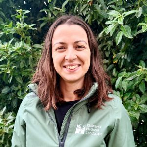 Headshot of a woman in a Chilterns National Landscape shirt in front of green foliage.