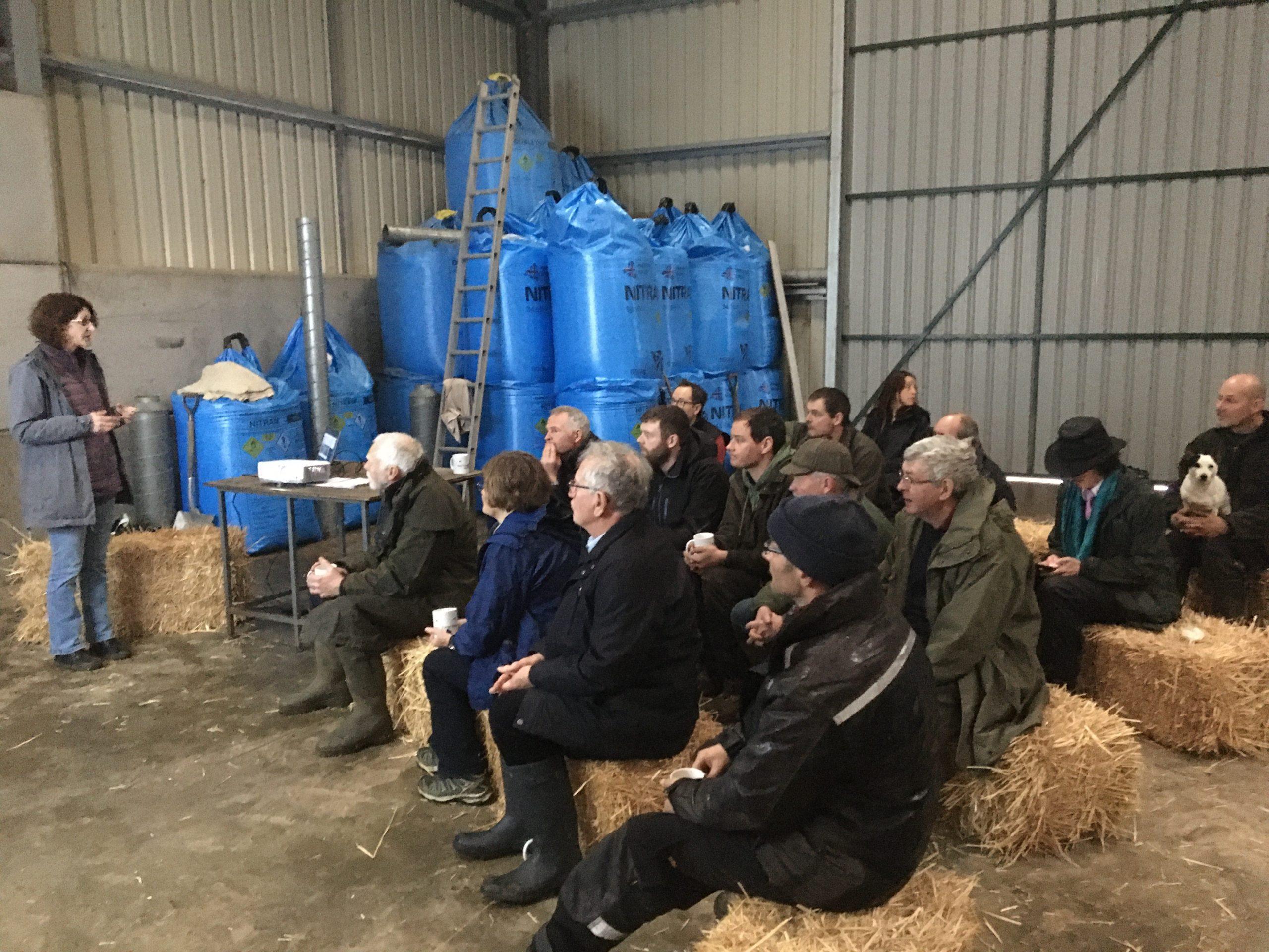 A group of farmers sitting on straw bales in a barn listening to a woman giving a presentation.