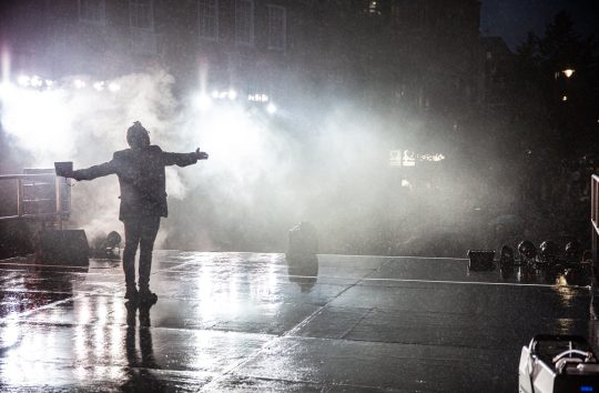 A dramatic image showing the silhouette of a man with his arms outstretched facing away from the audience with his back to the camera. He is performing on an empty stage with lights and smoke in the background.
