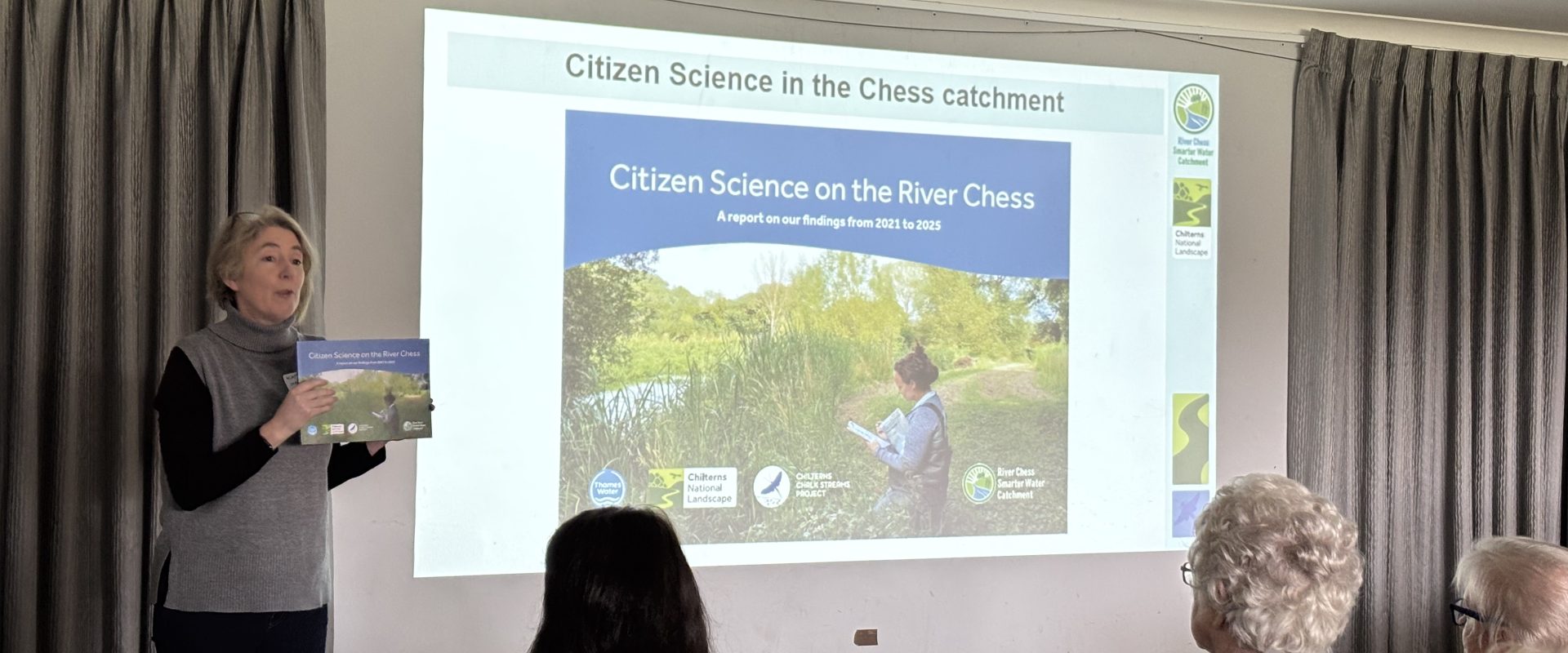 A woman stands in front of an audience with a presentation screen behind her. She is holding a paper booklet.