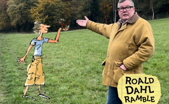 A man in a beige coat stands in a field and points at an illustrated boy in cargo pants and a blue t-shirt.