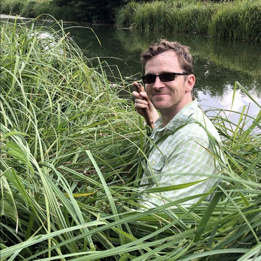 A man with short brown hair wearing sunglasses stands in a reed bed alongside a river.