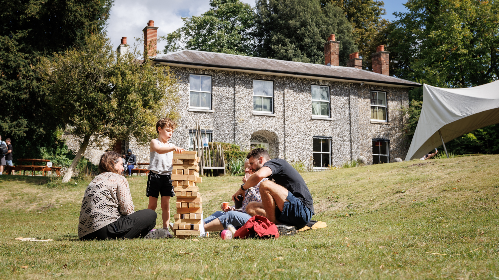 A couple with young children play with a giant Jenga set on the lawn in front of a stone building.