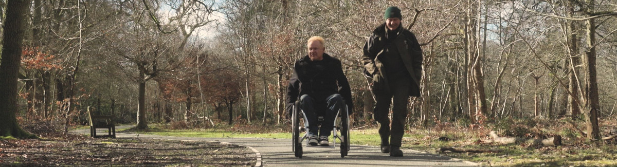 Two men on a path through a forest. One is in a wheelchair while the other walks beside him.
