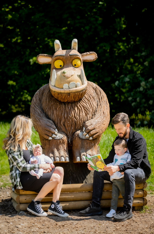 woman with baby sits by gruffalo statue while man reads to toddler from gruffalo book