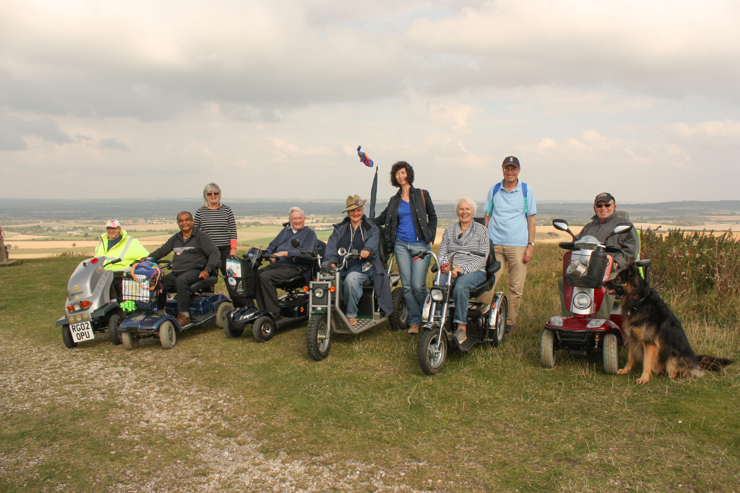 A group of 6 people in mobility scooters, 2 people standing and a German shepherd dog pose for a photo at the top of a hill.