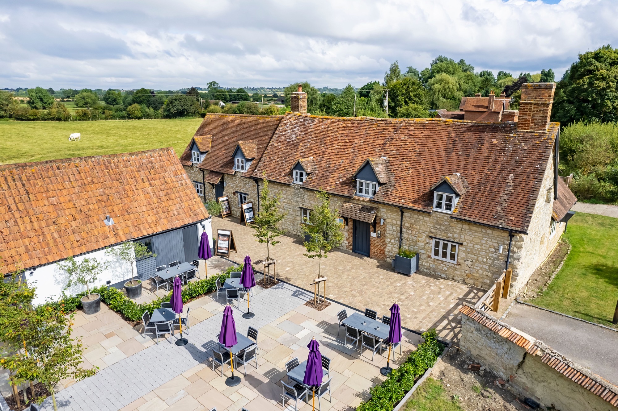 Aerial photo of a pub. A patio with chairs, tables and purple umbrellas with a old country inn style building behind it.