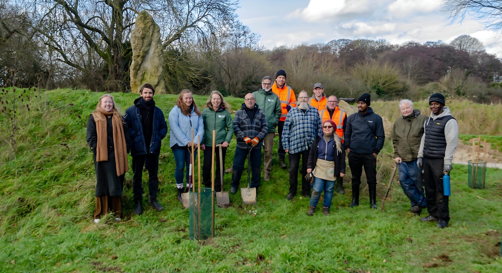 A group of about 15 people stand in a field. They have spades.