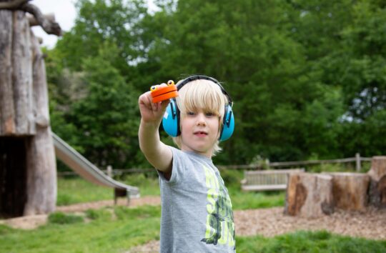 A young child with blond hair wearing a grey tshirt and bright blue ear defenders holds up an orange toy.