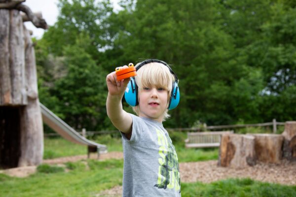 A young child with blond hair wearing a grey tshirt and bright blue ear defenders holds up an orange toy.