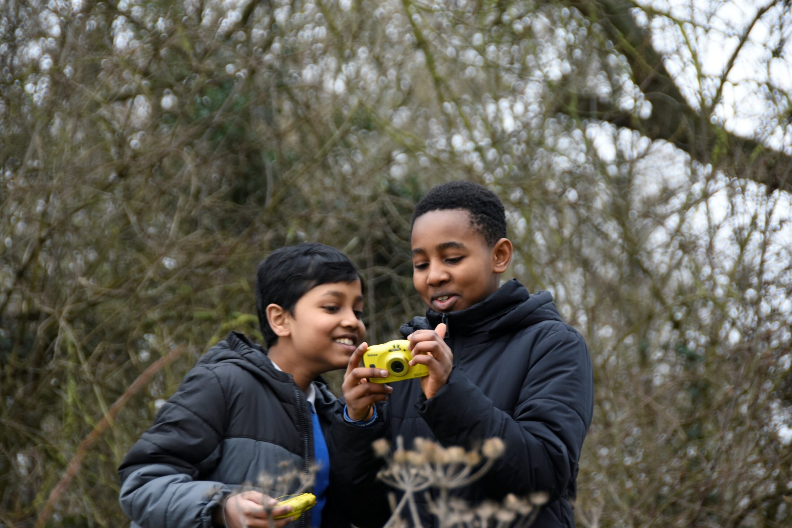 Two young boys stand in front of a tree, one is holding a digital camera and both are looking at the screen and smiling.