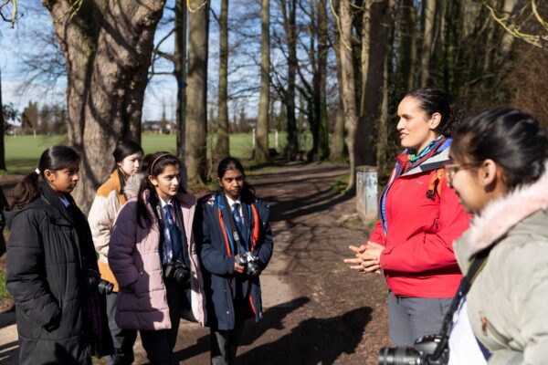 Five teenage students talk to a women in a red coat while in a woodland. The students have DSLR cameras.