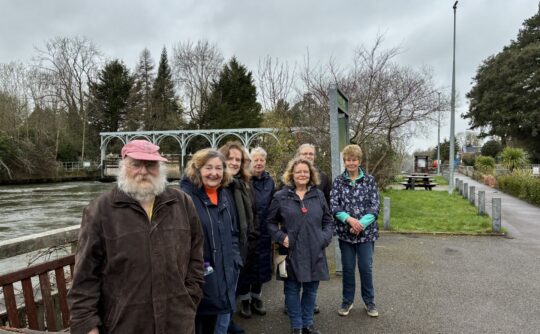 A group of 6 older people pose for a photo on a footpath alongside a river.