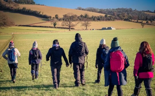A group of 7 adults walk across a field. They are wearing outdoor gear and walking away from the camera.