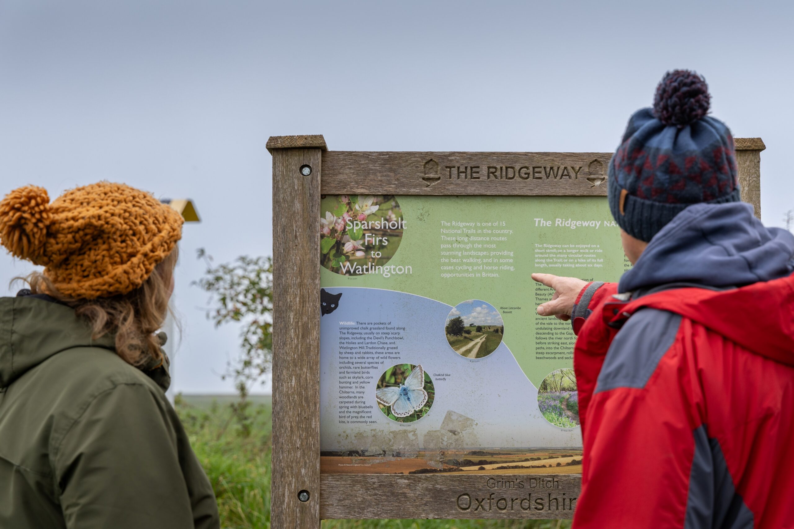 Two people in waterproof coats and knitted hats stop to read a sign about the ridgeway.