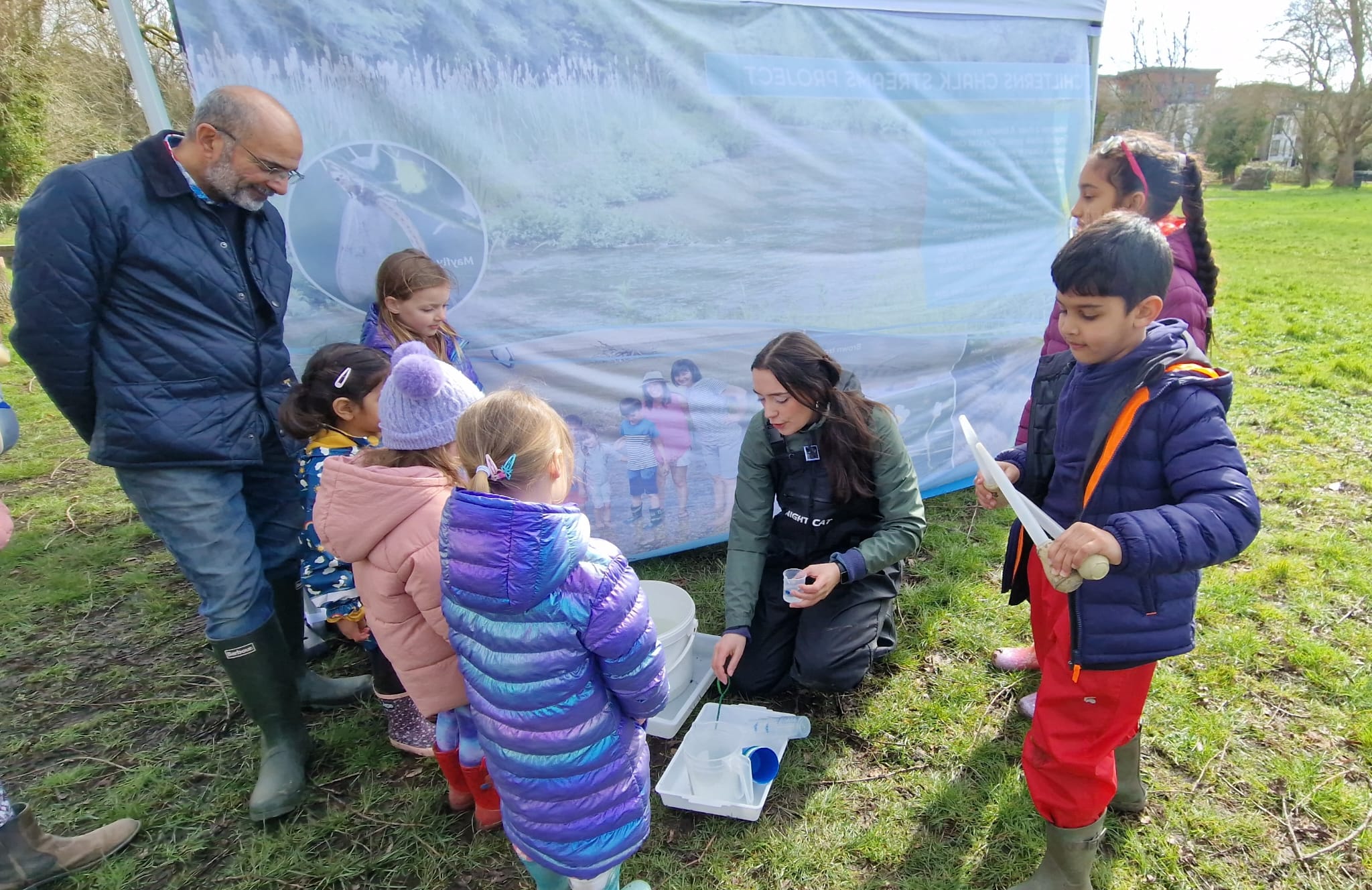Six young children gather round a woman as she kneels in front of a white tray full of river water and invertebrates.
