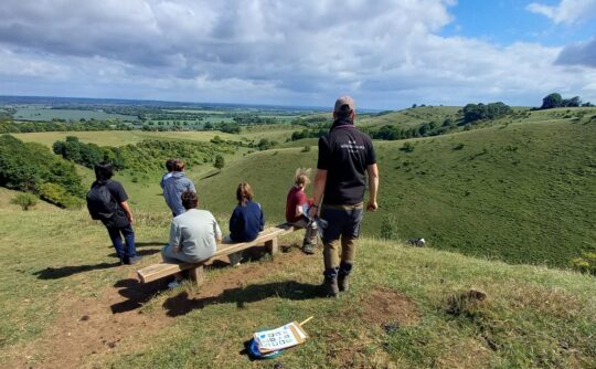 A group of 6 people sit on a bench at the top of a hill and look out at the view.