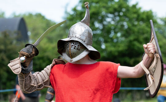 A man dressed as a Roman gladiator in a red shirt, metal hat with a shield.