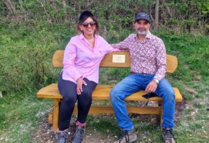 A woman in a pink shirt and jeans sits on a bench next to a man in a red paisley shirt and jeans.