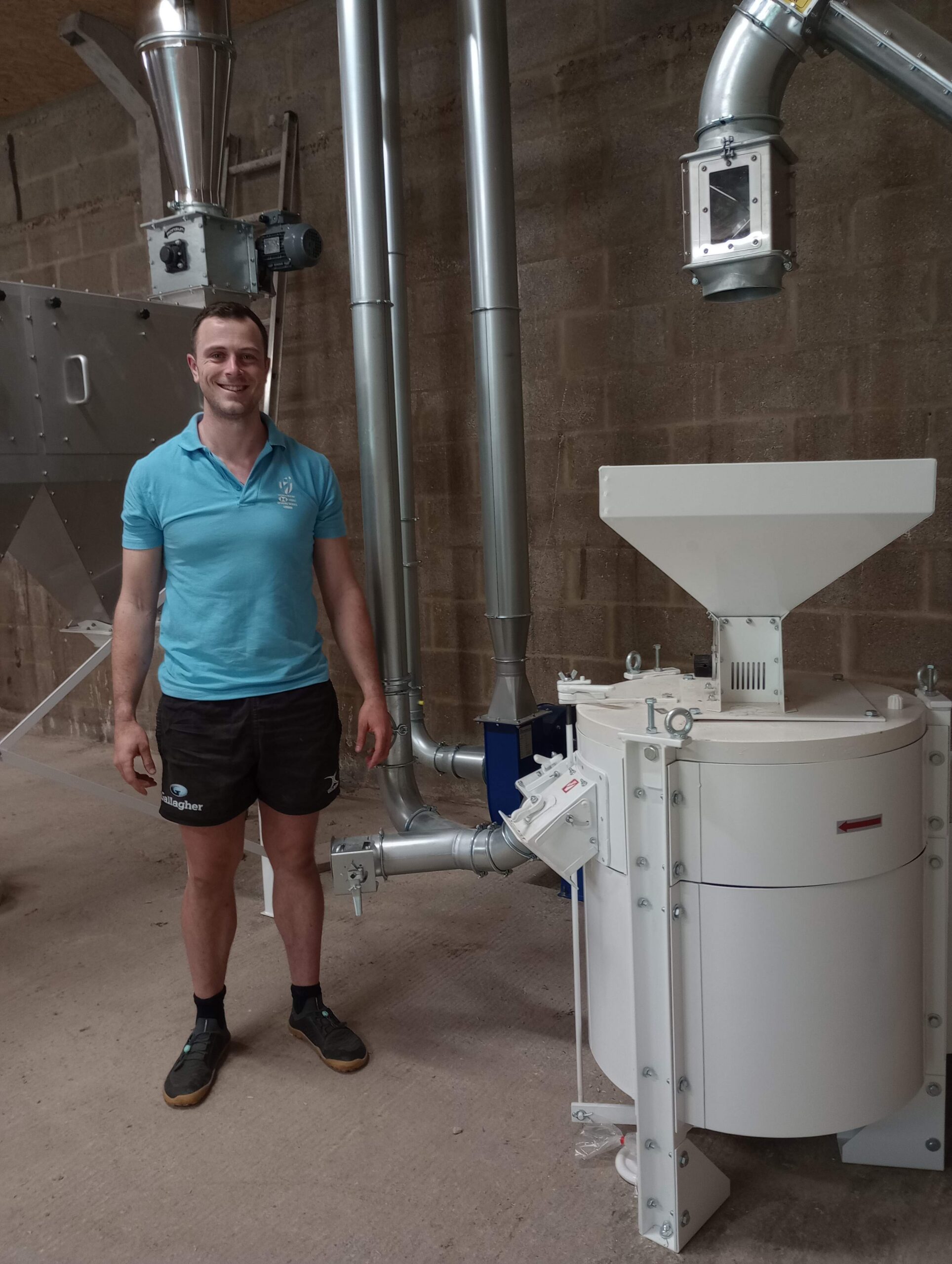 A man in shorts and a tshirt stands in a barn next to a white grain still and silver pipes.