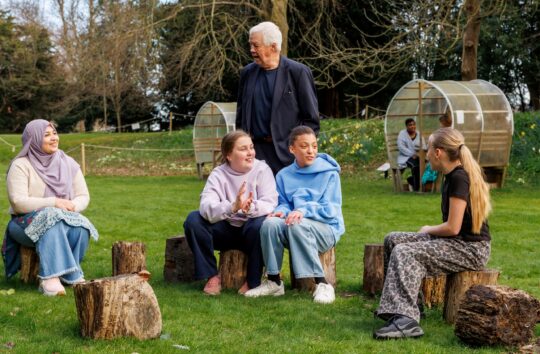 A group of four teenage girls sit on stumps in a garden chatting and laughing