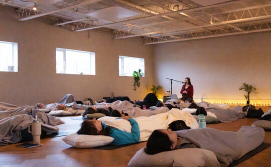 A room with a group of around 10 people lying on the floor with pillow and blankets and soft lighting.