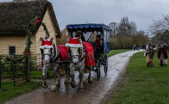 Two horses pulling a carriage with Christmas decorations along a track with a thatched cottage